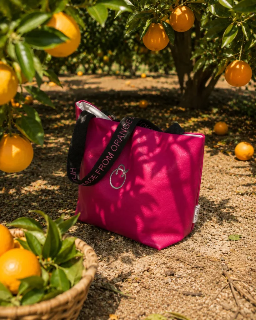 Pink JOY tote bag by Vestella resting on the ground in a Sicilian orange grove, beside a basket of freshly picked oranges