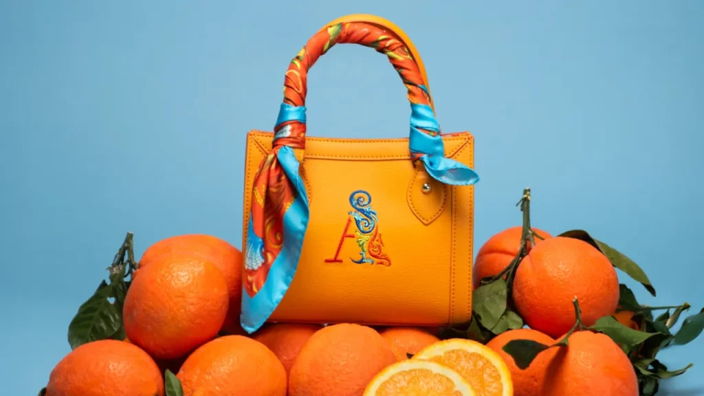 Orange Mini Bag by Ammia resting on fresh Sicilian oranges against a light blue background